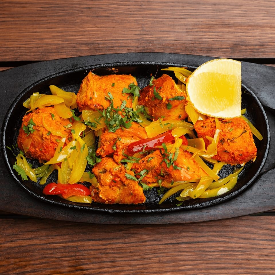 Indian chicken tikka with bell peppers, cilantro, and a lemon wedge on a black sizzling plate.