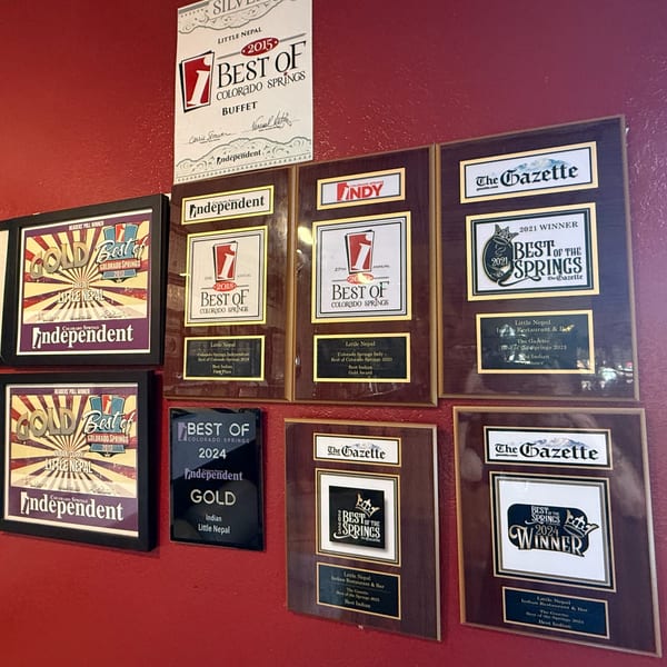 A red wall displays numerous framed awards and plaques for Little Nepal restaurant, including "Best of Colorado Springs".