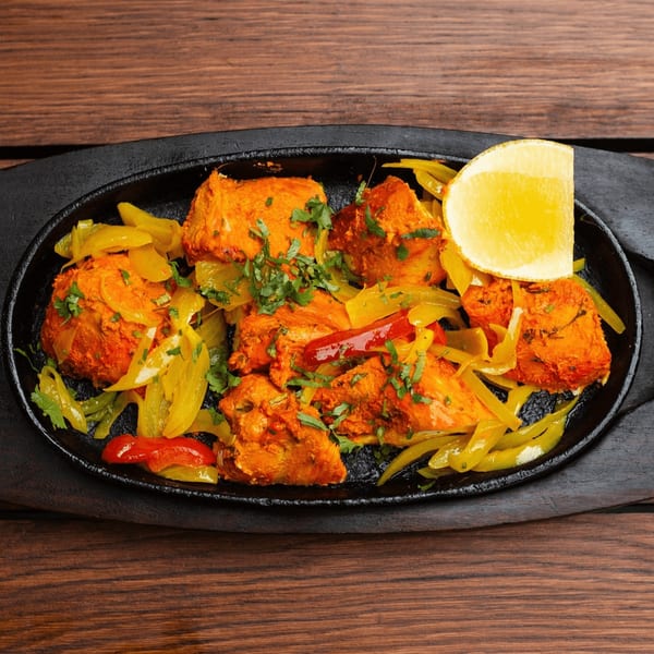 Indian chicken tikka with bell peppers, cilantro, and a lemon wedge on a black sizzling plate.